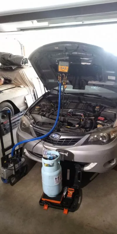 Subaru with hood up — A/C manifold gauge connected, refrigerant cylinder staged on scale in front of bumper