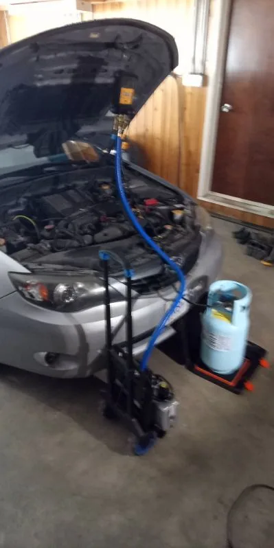Subaru in workshop with vacuum pump and refrigerant cylinder connected on scale during weighed A/C recharge